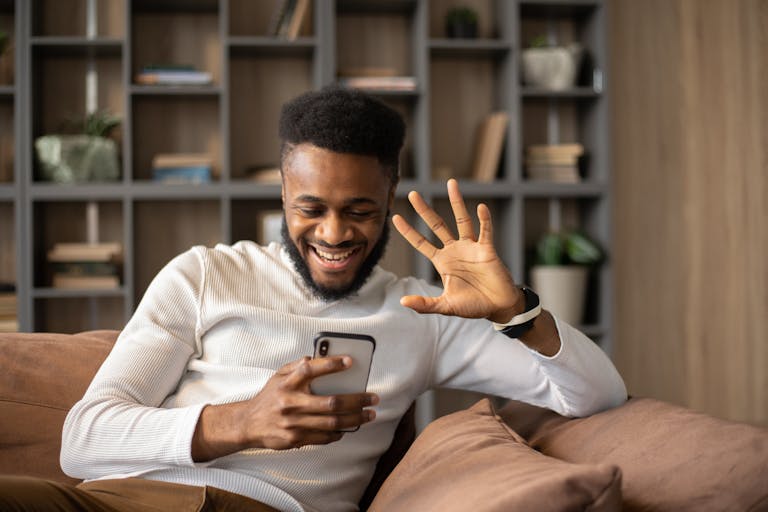 Smiling man relaxing on couch, waving during an engaging smartphone video call indoors.