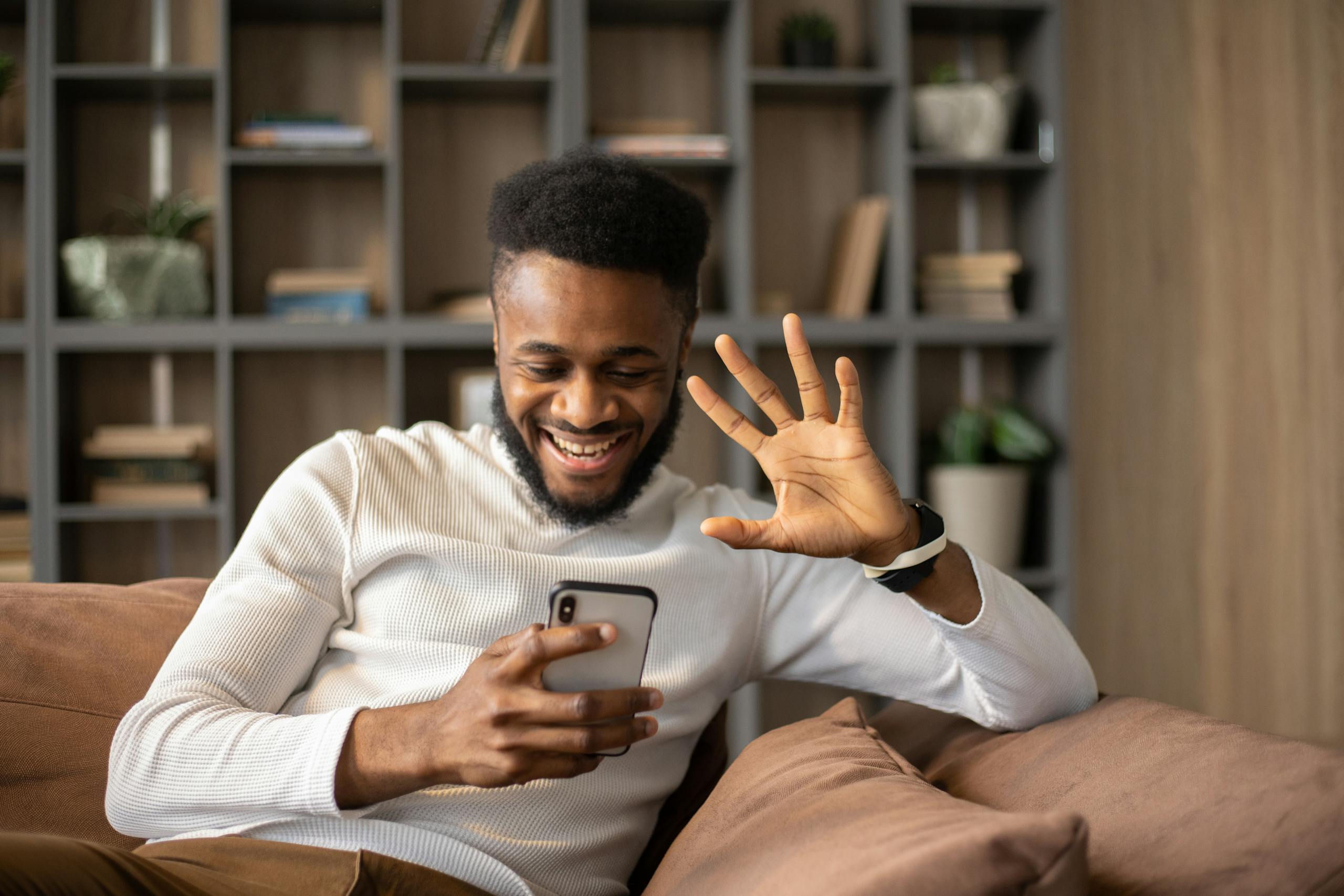 Smiling man relaxing on couch, waving during an engaging smartphone video call indoors.